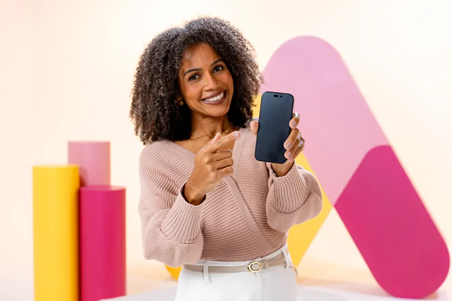 Mulher sorrindo apontando para a tela de um celular diante de um fundo colorido